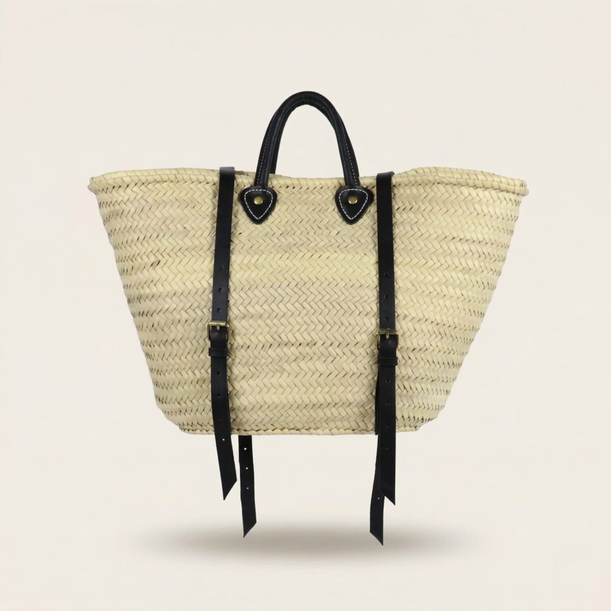 Handwoven Moroccan palm leaf straw market tote with braided leather top handles and convertible leather backpack straps with silver buckles on white surface