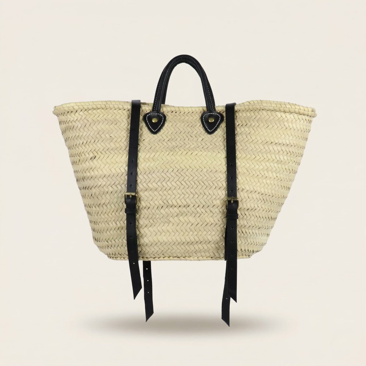 Handwoven Moroccan palm leaf straw market tote with braided leather top handles and convertible leather backpack straps with silver buckles on white surface
