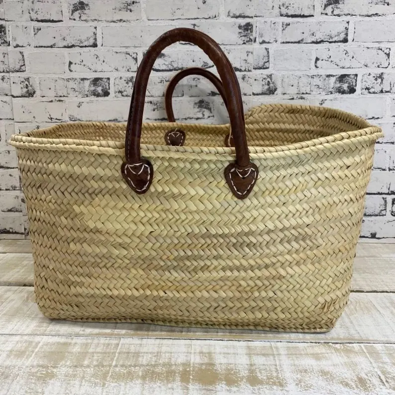 il_794xN.7593299542_t0ud Handmade Moroccan palm leaf straw market basket with short dark leather loop handles and heart-shaped anchor tabs, wide oval open-top silhouette, classic French market style