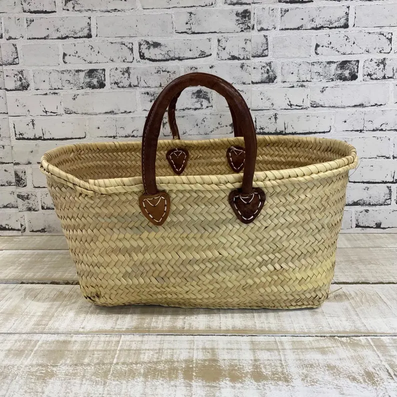 il_794xN.7593299552_qjus Handmade Moroccan palm leaf straw market basket with short dark leather loop handles and heart-shaped anchor tabs, wide oval open-top silhouette, classic French market style