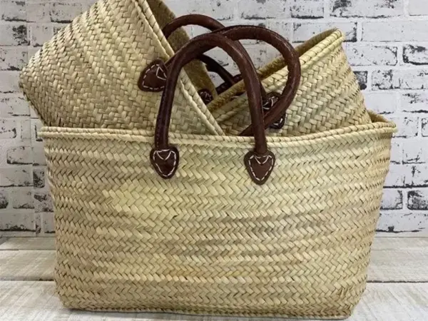 Handmade Moroccan palm leaf straw market basket with short dark leather loop handles and heart-shaped anchor tabs, wide oval open-top silhouette, classic French market style