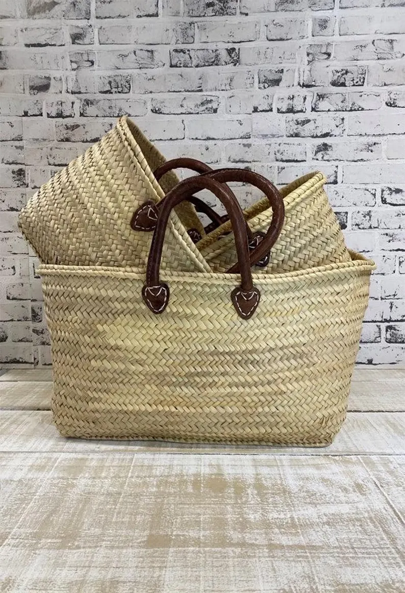 Handmade Moroccan palm leaf straw market basket with short dark leather loop handles and heart-shaped anchor tabs, wide oval open-top silhouette, classic French market style